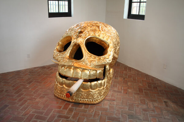 A large gilded skull, smoking a cigarette, sits on the floor of a tiled white room.