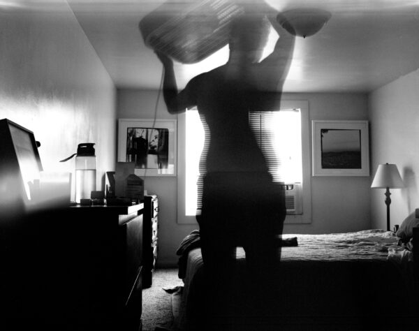 A photograph featuring the outline of a body, in a bedroom, holding something above their head.
