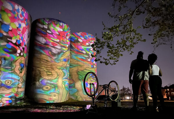 A night time photograph of two people watching a brightly colored abstract video work projected on three large cylinder structures.