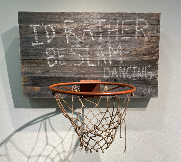A photograph of a sculpture by Chad Rea of a basketball hoop attached to a wooden backboard with text reading, "I'd Rather Be Slam-Dancing."