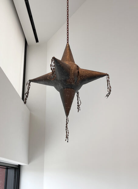 A photograph of a hanging metal sculpture by David Avalos, made to look like a traditional star-shaped piñata.