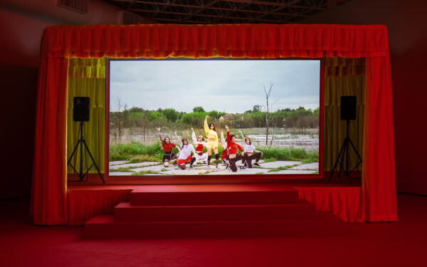 A photograph of a projected video on a large screen set on a red stage with red curtains around it.