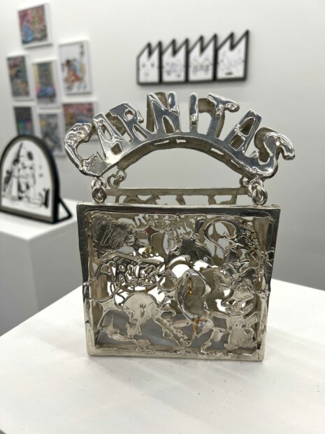 A silver art object sits on a pedestal. The piece looks like an open bag, with ornate metalwork. 