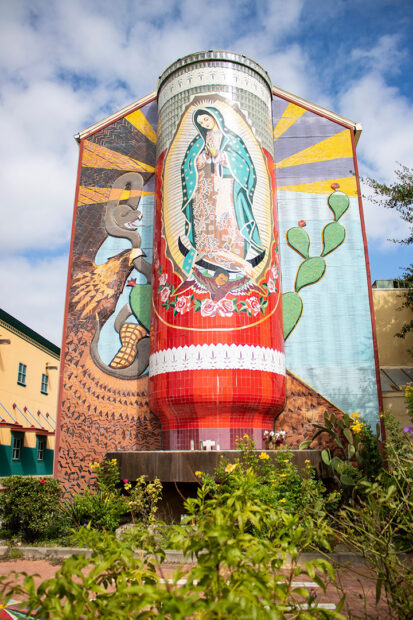 A photograph of a large mosaic mural featuring a building-size three-dimensional votive candle with the image of Our Lady of Guadalupe.