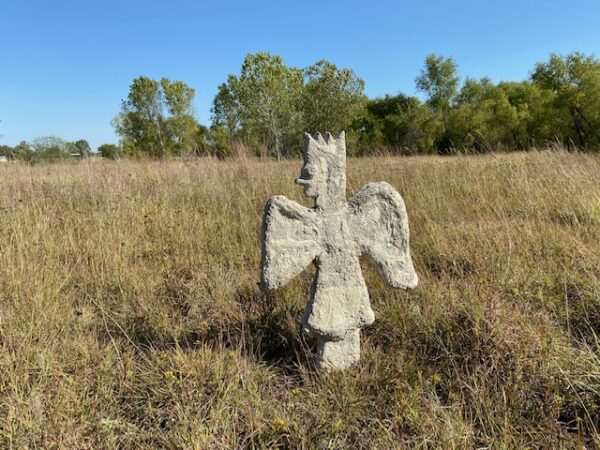A concrete sculpture by Joshua Goode of a winged figure with the profile of the cartoon character Bart Simpson. The sculpture is placed standing in a large empty field.
