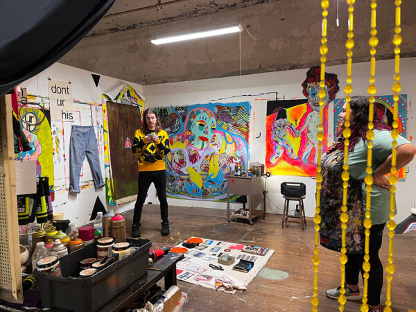 A photograph of two people standing in an artist's studio with bright stylized paintings in the background.
