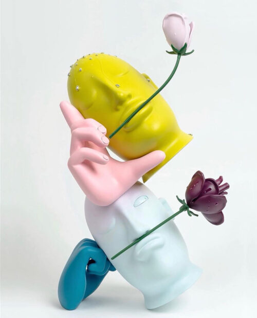 A sculpture by Mark Whalen featuring two heads and two hands stacked and balanced. The figures are painted pastel colors and each head holds a flower in or above its mouth.