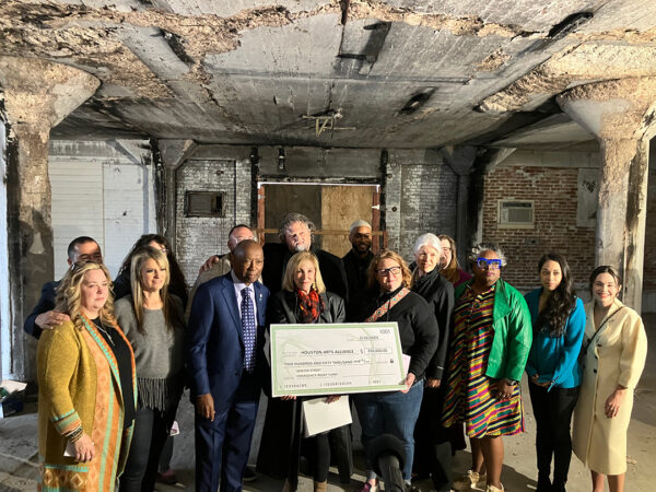 A crowd of people stand in a severely burned building and hold a large check, which documents a donation of $250,000.