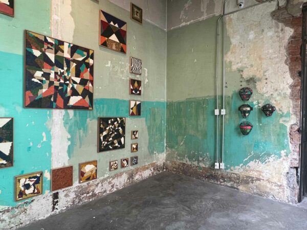 Many rectangular and square artworks hang on a turquoise wall. Opposite, on another wall, are small, mask-like objects.