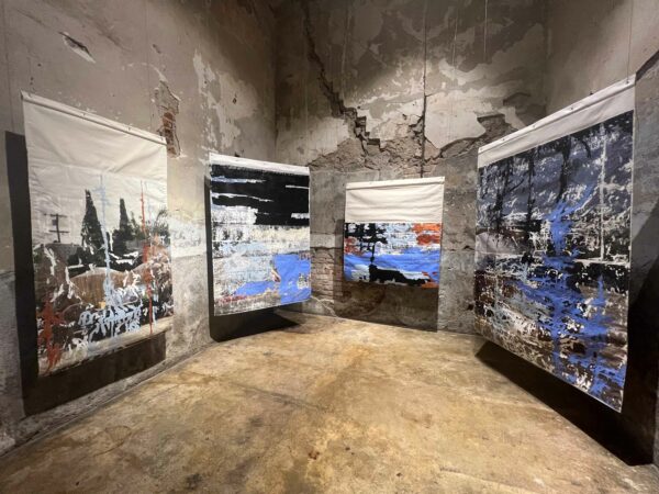 Large-scale collaged-looking artworks hang in a rough, concrete space.