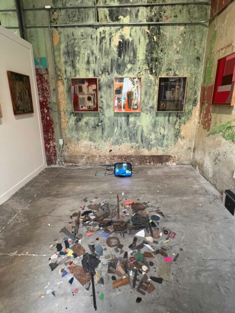 A circle of objects sits on a concrete floor, and paintings are on view on three walls.