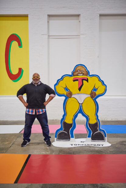 A photograph of Trenton Doyle Hancock standing next to a large cut-out figure of a super hero as part of an installation he created at MASS MoCA.