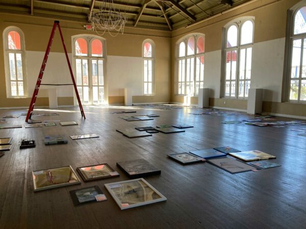 Framed artworks sit on a wooden floor of a windowed, yellow, light-filled room. A ladder sits in the distance.