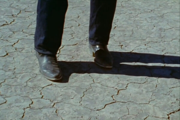 Video still showing a mans legs in cowboy boots standing on dry, cracked land