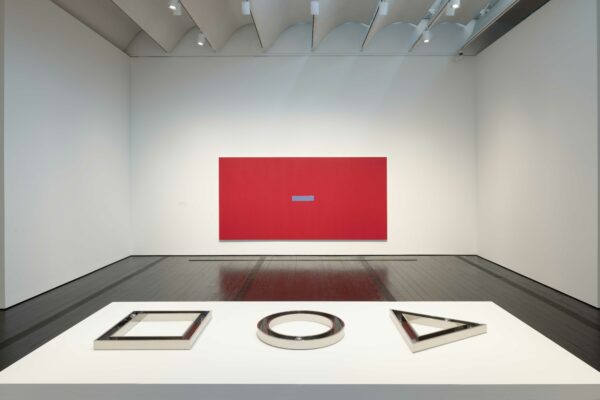 Installation view of a large red canvas with three works resting on top of a white plinth in the foreground
