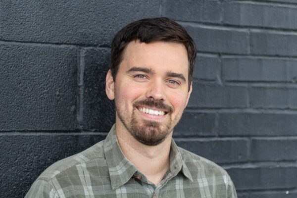 Un hombre joven sonriente de tez clara y barba corta posa frente a un muro de ladrillos negros.
