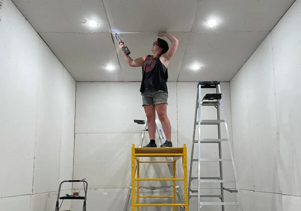 A photograph of a woman standing on scaffolding as she constructs the ceiling of an artist studio space.