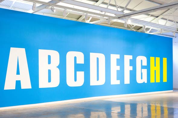 An installation image of a text-based mural by Kay Rosen featuring the first nine letters of the alphabet with the letters "H" and "I" rendered in bright yellow.
