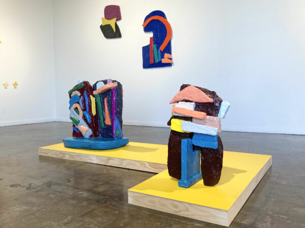 Installation view of two sculptures on a yellow, L-shaped plinth and two dimensional works hanging on a wall behind