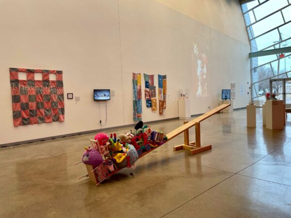 Installation view with two dimensional works hanging on a wall, and a seesaw in the middle of the space