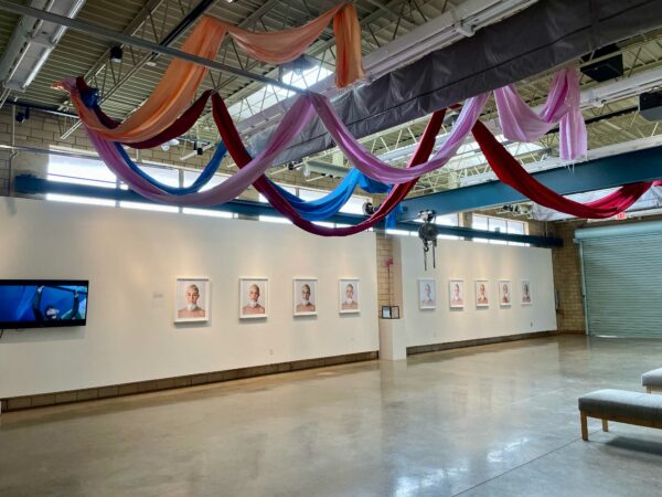 Installation view of photos hanging horizontally on a white wall and colorful fabric banners in the rafters