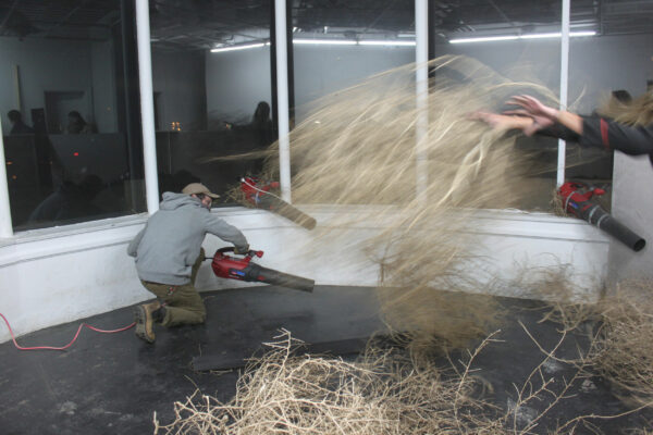 Artists using leaf blowers to move tumbleweed in a white gallery space