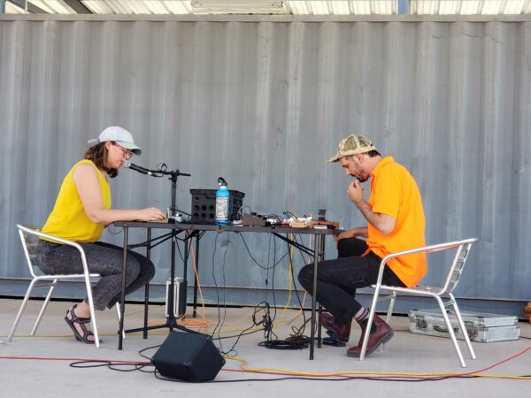 Two people in yellow and black playing music from synthesizers across a table