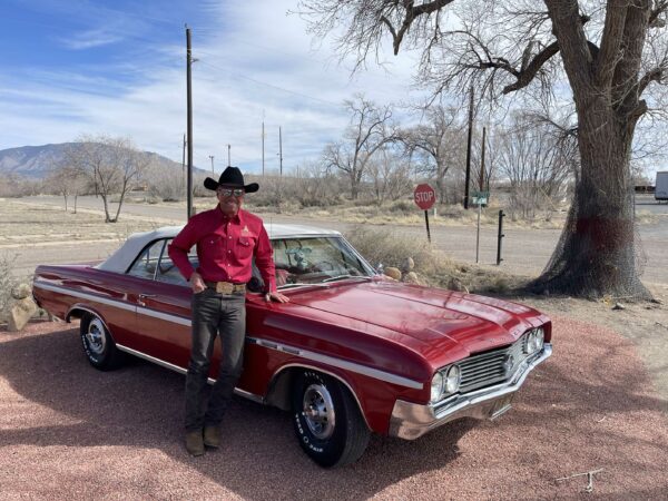 Photo of a man wearing a black stetson and a red pearl snap button down standing next to a red skylark car