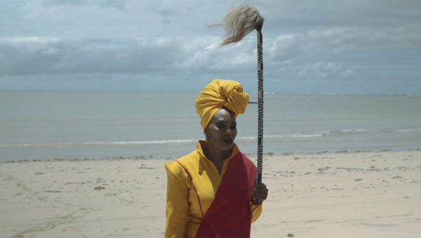 A still image from a video by Lhola Amira. The image shows a Black woman standing on a beach and holding a staff.