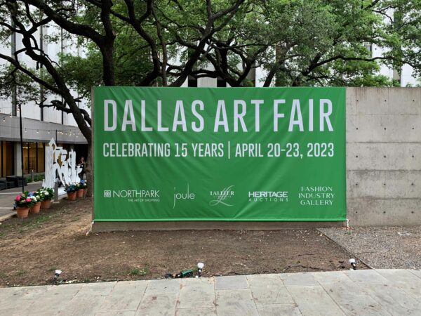 A large green banner reads DALLAS ART FAIR. It is hung on a concrete wall in a wooded area.