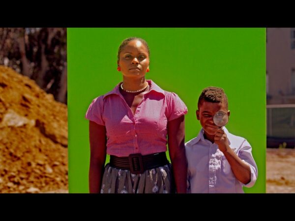 A still image from a video work by Cauleen Smith. The image depicts a young Black boy holding a magnifying glass up to his eye and standing next to a Black woman in front of a green screen. 