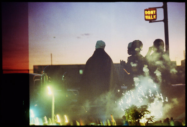 A photograph taken around dusk of a group of silhouetted men standing near a crosswalk. Photograph by Ming Smith.