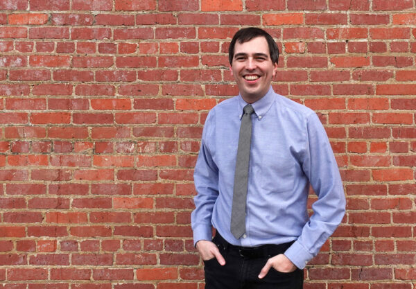 A photograph of curator and cultural worker Caleb Bell standing in front of a red brick wall.