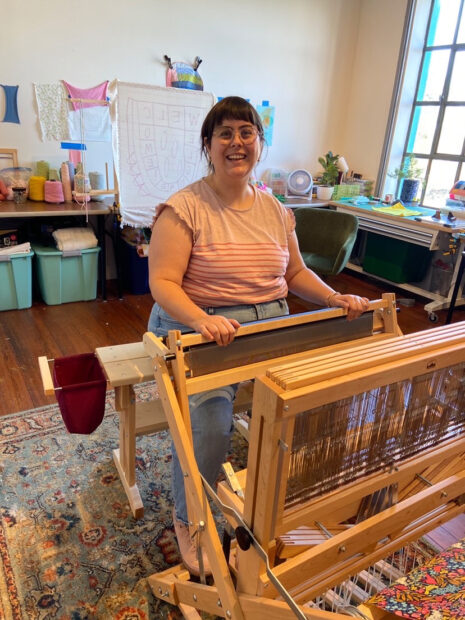A photograph of artist Chelsea Clarke seated at a large loom.
