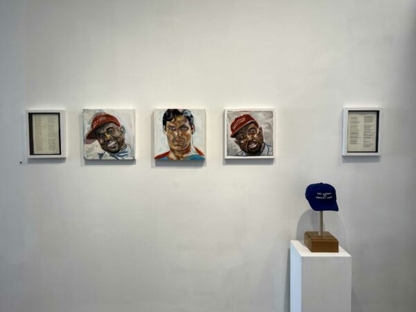 Portraits of Kanye West and superman and a blue baseball cap on a pedestal