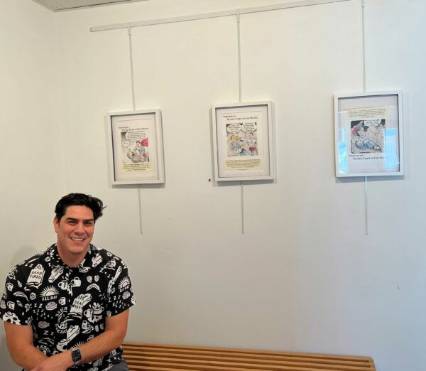 The artist sitting in front of three drawings hanging on the wall