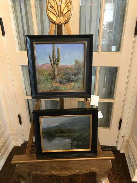 Two paintings stacked on an easel, the top of a desert landscape, and the bottom a river landscape