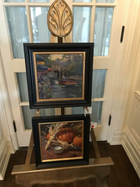 Two paintings stacked on top of each other on an easel, the top a view of the riverwalk, and the bottom a still life with pumpkins