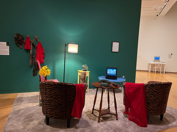 A photograph of an installation and digital video work by Lynn Hershman Leeson featuring a video displayed on TV within a small space set up like a living room.