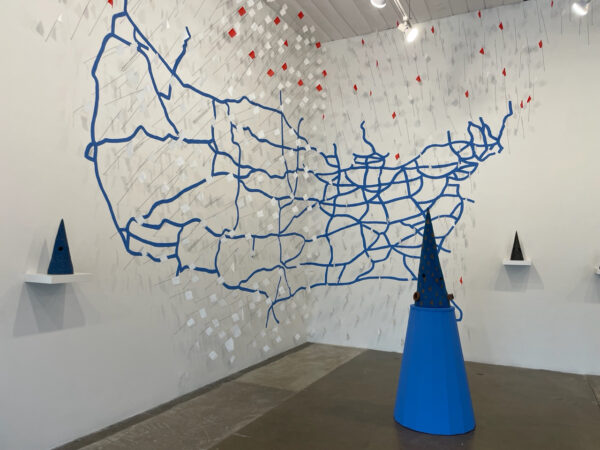 Installation view of a blue tape map of the united states with cone shaped ceramic sculptures on pedestals