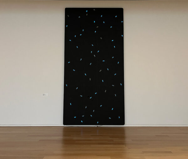 An installation photo of a large vertical artwork featuring 61 blue LED counters randomly spread across a black wooden panel. Artwork by Tatsuo Miyajima.