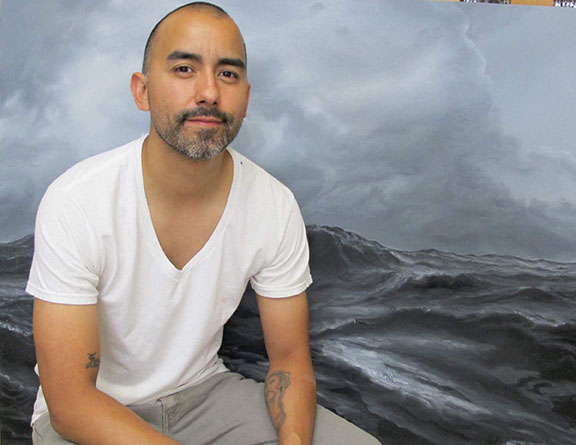 A photograph of painter Vincent Valdez sitting in front of a painted canvas.