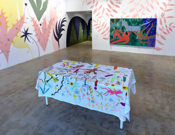 Installation view of pastel floral paintings on a wall, a landscape scene and a table with an embroidered white tablecloth