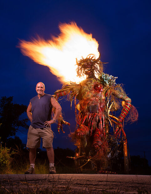 A nighttime photograph of Mark "Scrapdaddy" Bradford standing next to his large kinetic sculpture, with flames coming from the top.