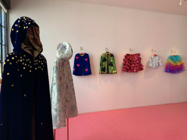 Multiple fabric capes, made of sequins and various other decorative materials, hang in a gallery space.