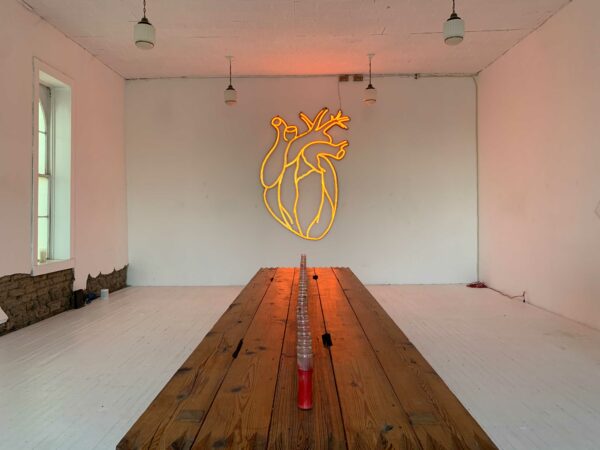 There is a table running down the middle of a clean, white gallery space. In the distance is a large LED heart.