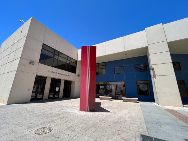 Vista exterior de un edificio de dos pisos en forma de L color concreto y con ventanas oscuras. Los muros a la sombra del lado derecho son de color azul eléctrico y en el centro de la placita frontal se alza verticalmente una escultura geométrica roja. 