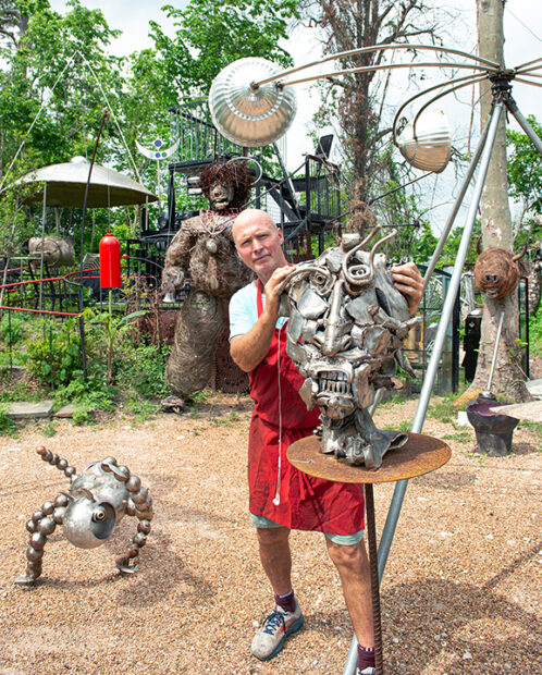 A photograph of artist Mark "Scrapdaddy" Bradford standing with an array of metal sculptures.