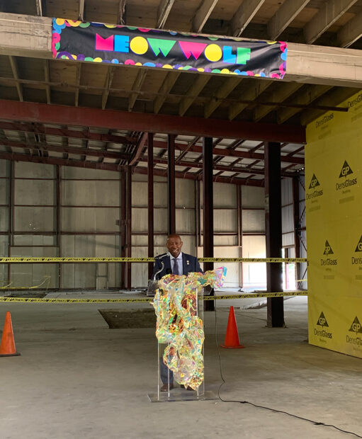 A photograph of Houston's Mayor Sylvester Turner standing at a podium inside of an empty warehouse with a banner overhead that reads, "Meow Wolf."
