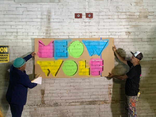 A photograph of two men holding a large stencil used to spray paint the Meow Wolf logo on a white brick wall.